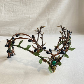 Front view of a bronze-tone woodland antler tiara with a green centre accent and dark buds threaded through leafy branches.