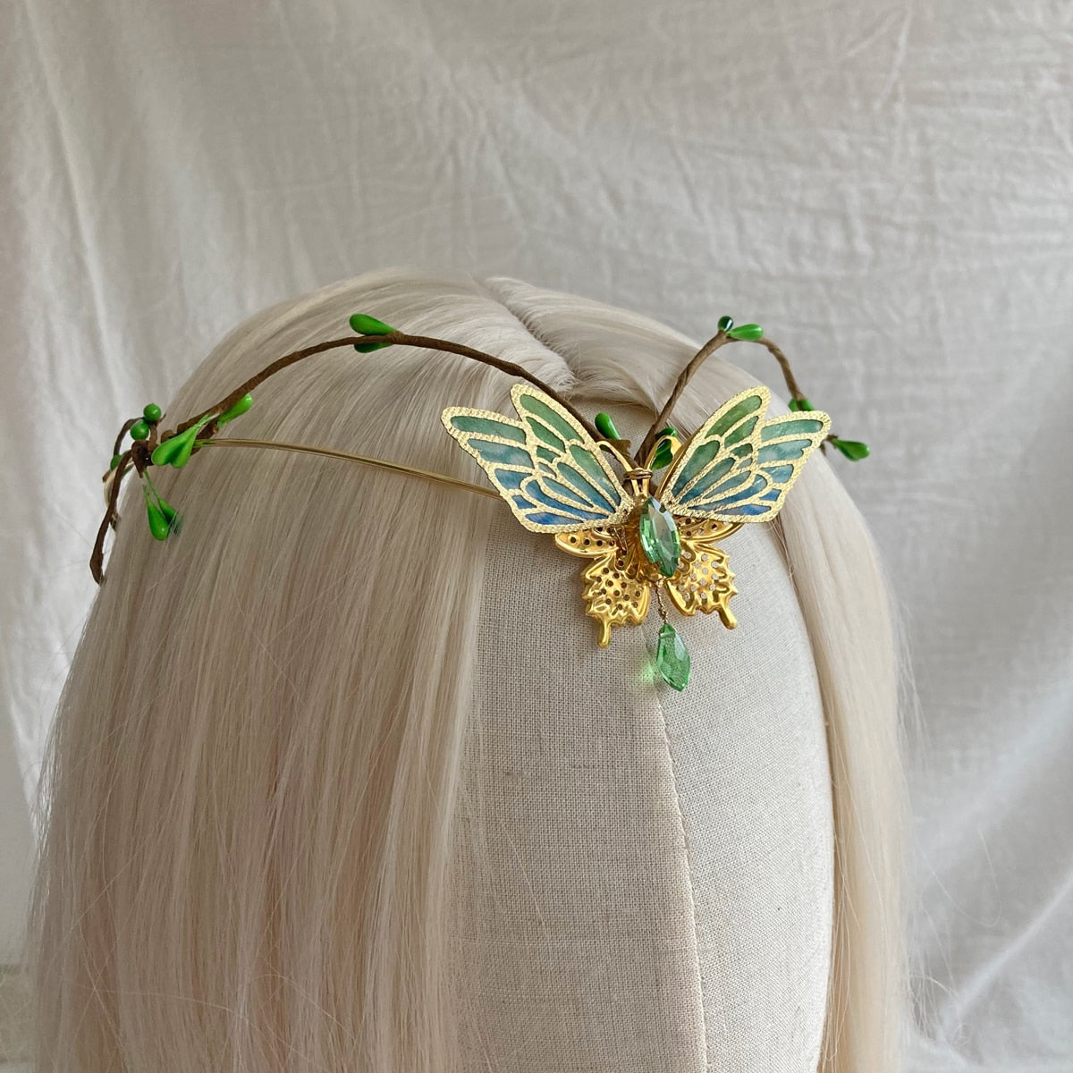 Front view of a gold-tone butterfly tiara with green-blue wings, small leaf buds and a central green drop.