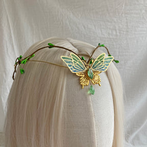 Front view of a gold-tone butterfly tiara with green-blue wings, small leaf buds and a central green drop.