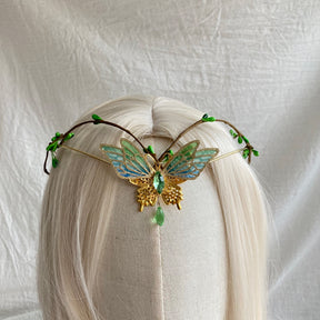Gold-tone butterfly tiara with green-blue openwork wings, tiny green buds and a green hanging drop on pale hair.