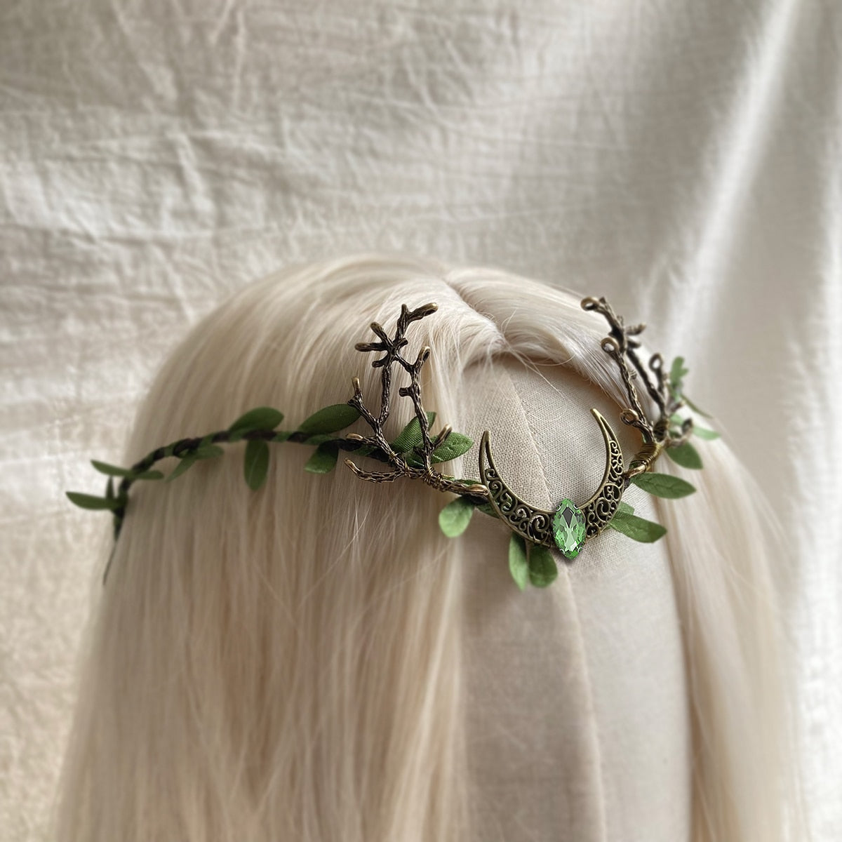 Bronze-tone crescent-and-antler tiara with green leaves and a small green teardrop at the centre, shown across pale hair.