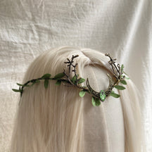 Bronze-tone crescent-and-antler tiara with green leaves and a small green teardrop at the centre, shown across pale hair.