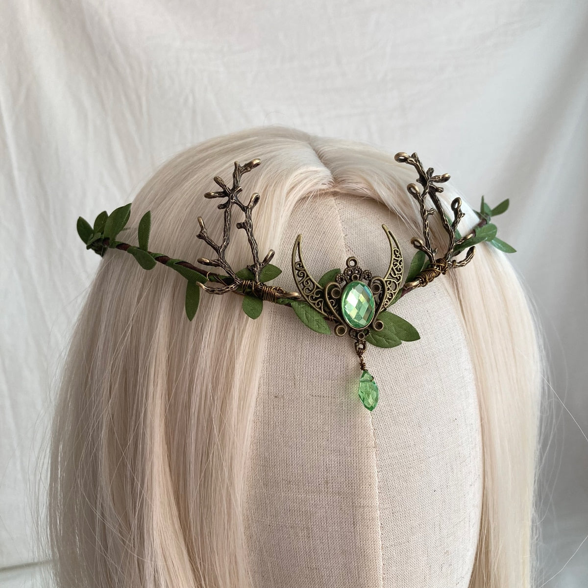 Angled view of a brown branch tiara with green leaves and a crescent framing a pale green oval accent and drop.