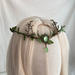 Angled view of a brown branch tiara with green leaves and a crescent framing a pale green oval accent and drop.