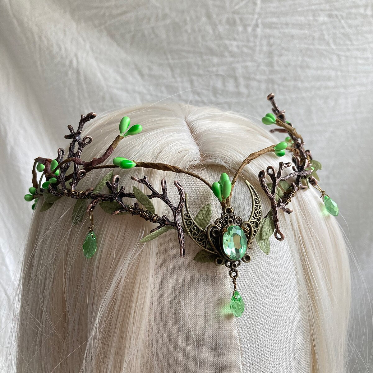 A pale green oval centre and drop hang from a crescent on a leafy antler tiara, photographed close against pale hair.