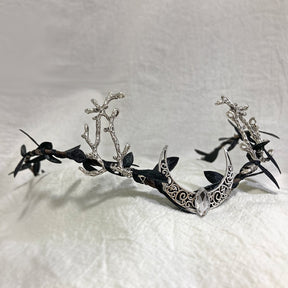 Silver-tone openwork crescent tiara with branching antlers, dark leaves and a clear drop at the centre.