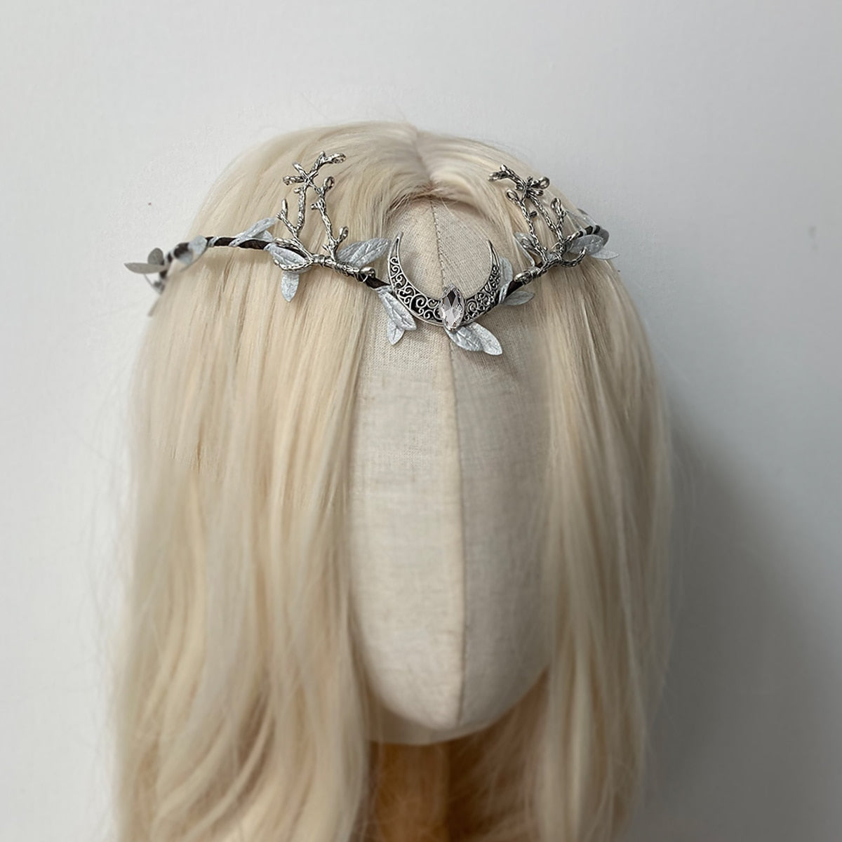 Silver-tone crescent and antler tiara on pale hair, with light leaf accents and a clear central drop.