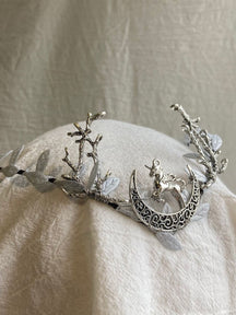 The silver-tone unicorn-and-crescent tiara rests on pale fabric, with branch-shaped antlers and light leaves rising on either side.