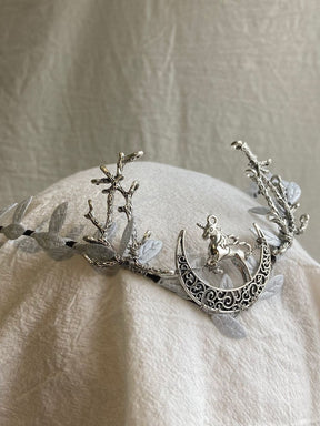 The silver-tone unicorn-and-crescent tiara rests on pale fabric, with branch-shaped antlers and light leaves rising on either side.