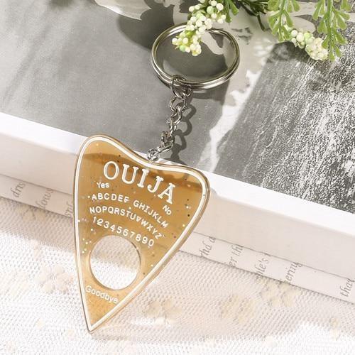 Translucent amber planchette keychain with glitter, white lettering and a circular opening, displayed below a silver-tone keyring beside small white flowers.