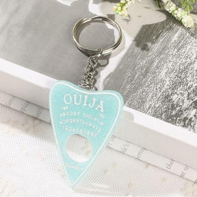 Pale aqua glitter planchette resin keychain with white alphabet and number details, resting on lace at the edge of an open book.