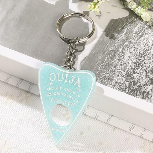 Pale aqua glitter planchette resin keychain with white alphabet and number details, resting on lace at the edge of an open book.