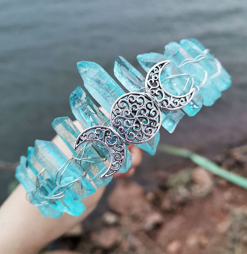 Angled view of a aqua crystal point crown with a silver-tone triple moon emblem and wire-wrapped band.