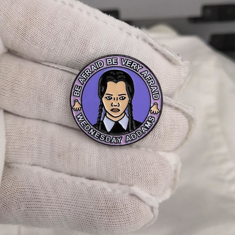 Purple Be Very Afraid portrait pin held against a dark glove, showing the pale face, black braids and surrounding lettering.