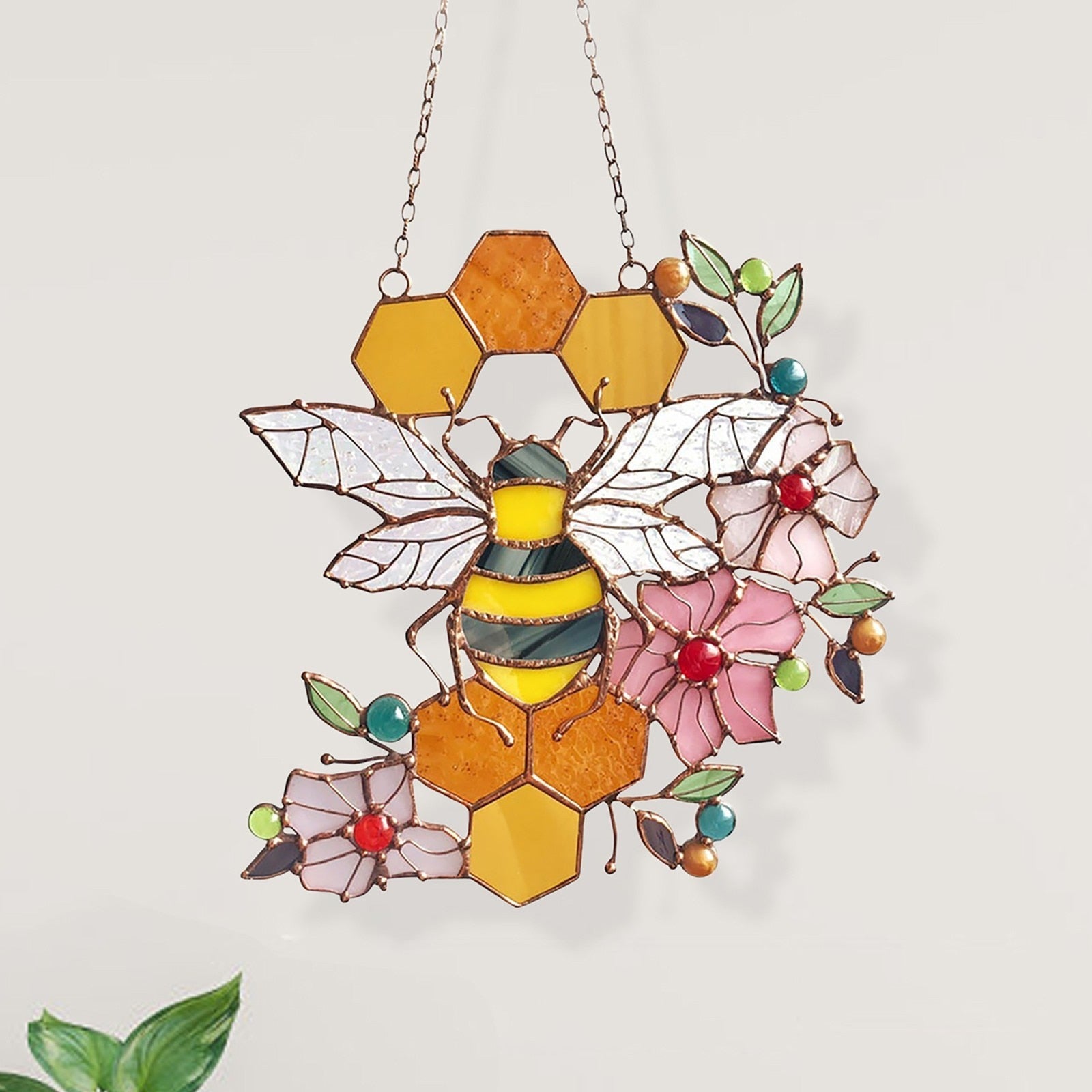 Bee and honeycomb stained glass hanging shown from the front beside a plant.