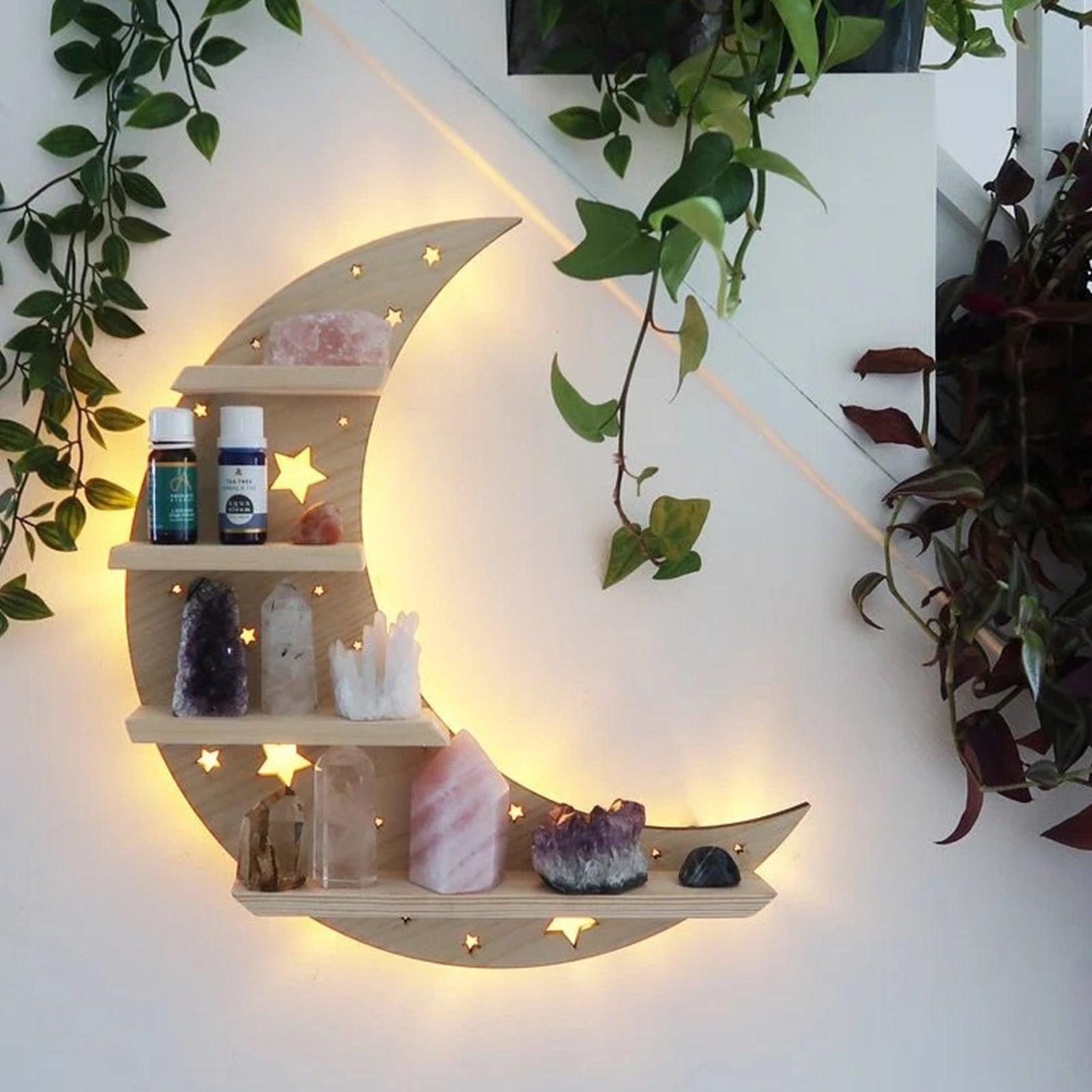Beige crescent moon shelf glowing against a pale wall, with crystals and small bottles arranged on its ledges and trailing greenery beside it.