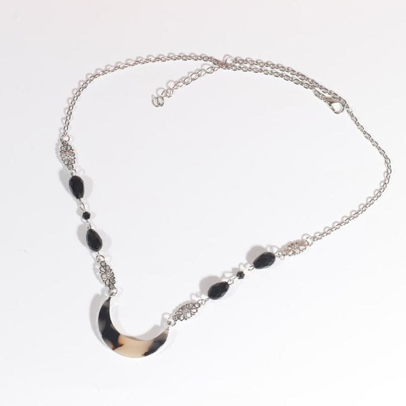 Silver-tone head chain with a beige-and-black tortoiseshell-style acrylic crescent and black teardrop beads, arranged in an oval on a pale background with the clasp and chain visible.