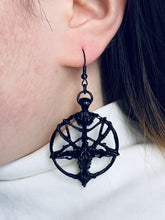 Black Baphomet pentagram drop earring worn beside dark hair, with an open circular frame and horned-goat detail above a white top.