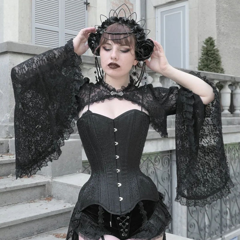 Model holding both hands beside a black headpiece, spreading the ruffled bell sleeves of a black lace overlay over a corset.