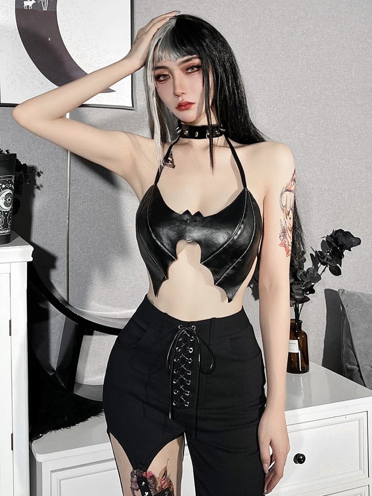 Black bat wing bralette worn with lace-up trousers and a choker beside white furniture. The model raises one arm, leaving the halter shape clear.