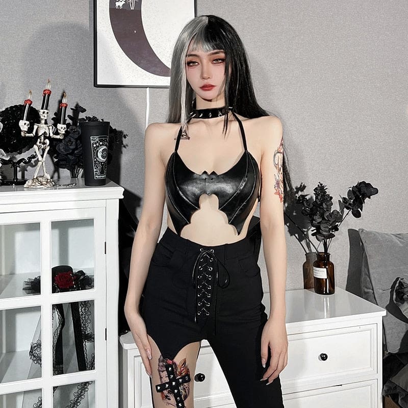 Black bat bralette and lace-up trousers worn in a gothic bedroom setting with dark flowers and pale furniture. The cropped wing shape exposes the waist.