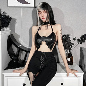 Model seated on white furniture in a glossy black bat bralette and lace-up trousers, showing the wing-shaped front against an open midriff.
