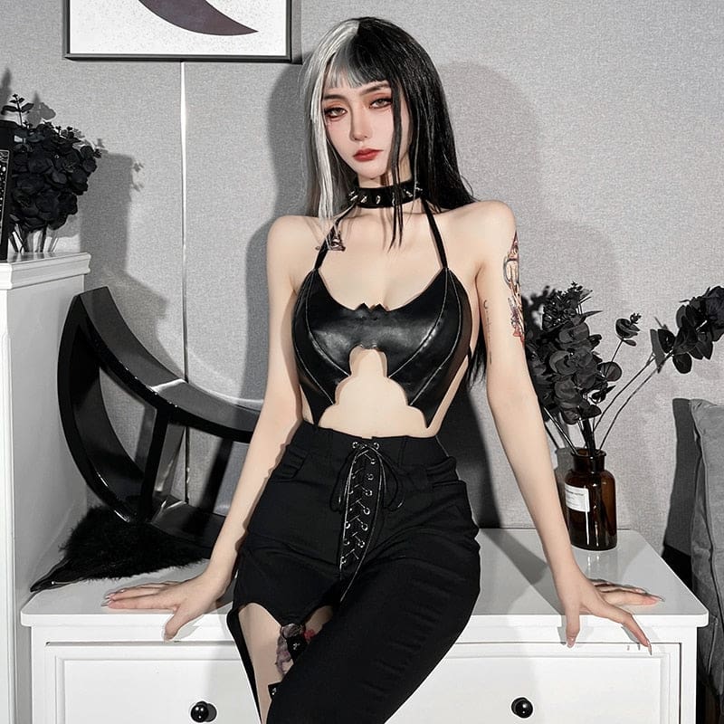 Model seated on white furniture in a glossy black bat bralette and lace-up trousers, showing the wing-shaped front against an open midriff.