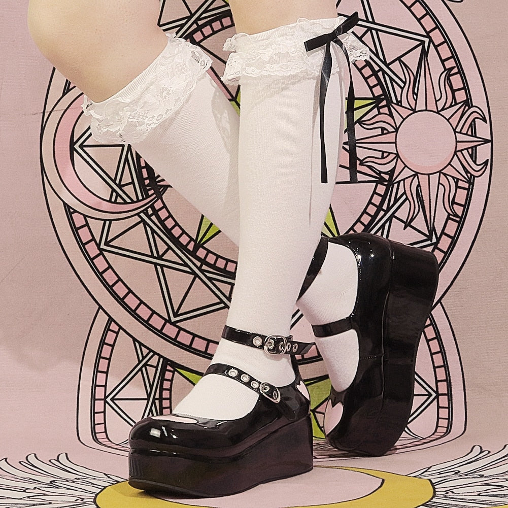 Glossy black heart platform Mary Janes styled with white knee-high socks and bows, photographed from the side against a pink illustrated backdrop.