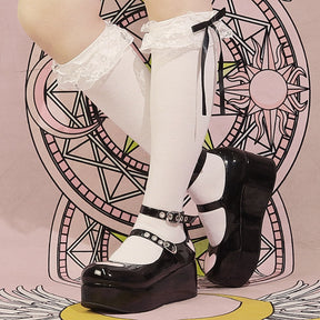 Glossy black heart platform Mary Janes styled with white knee-high socks and bows, photographed from the side against a pink illustrated backdrop.