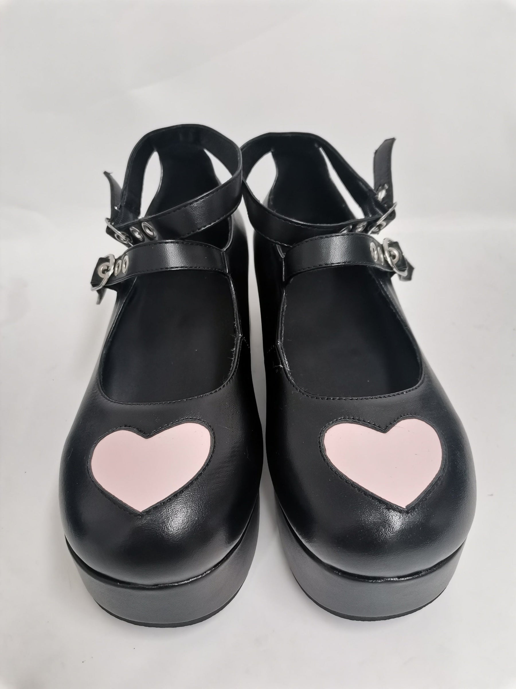Pair of matte black big-heart platform Mary Janes photographed from above, with large pink toe hearts and crossed buckle straps.