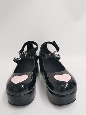 Pair of glossy black heart platform Mary Janes on a pale background, showing pink toe hearts, reflective uppers and buckle straps.