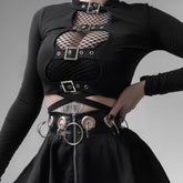 Black long sleeve buckle crop top layered over fishnet, with four silver-tone buckles framing the open front above a belted black skirt.