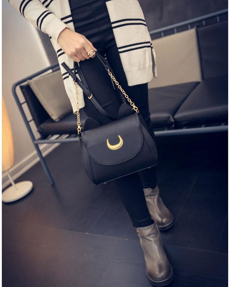 Black moon bag held by its chain section in a standing outfit photo, showing the compact front shape beside dark trousers.