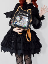 Black cat-ear display backpack with golden-orange trim and wide bat wings, held over a black ruffled outfit with pins visible through the clear front.