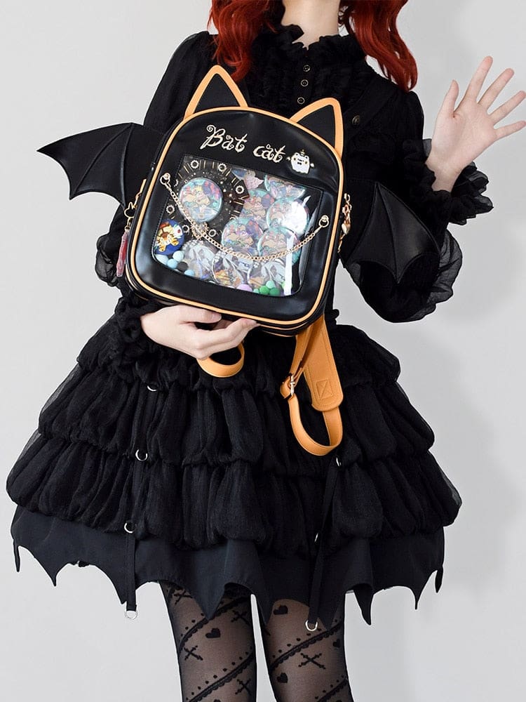 Black cat-ear display backpack with golden-orange trim and wide bat wings, held over a black ruffled outfit with pins visible through the clear front.