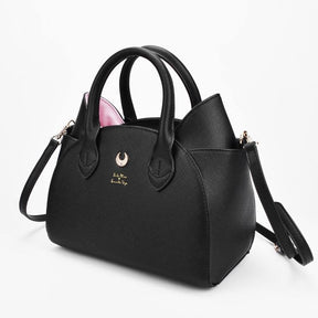 Angled view of a textured black cat-ear handbag with pink ear panels, curved handles and a gold-tone crescent, showing the depth of the side panel.