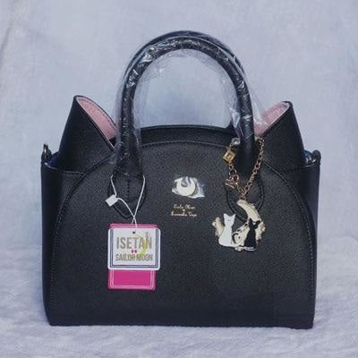 Black cat-ear handbag photographed with protective handle wrapping, retail tags and a hanging cat-shaped charm. Pink ears and a gold-tone crescent decorate the front.