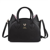 Black cat-ear handbag with pink inner ears, twin handles and a gold-tone crescent on the front. The long black shoulder strap extends beside the bag against white.
