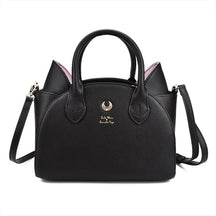 Black cat-ear handbag with pink inner ears, twin handles and a gold-tone crescent on the front. The long black shoulder strap extends beside the bag against white.