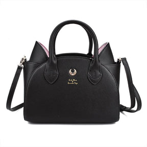 Black cat-ear handbag with pink inner ears, twin handles and a gold-tone crescent on the front. The long black shoulder strap extends beside the bag against white.