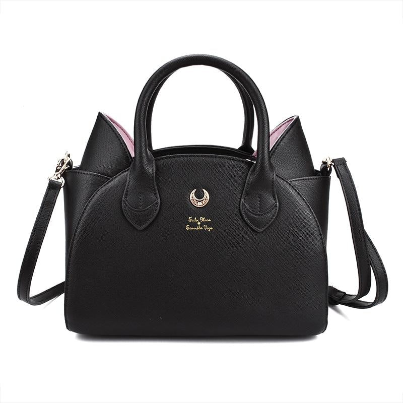 Black cat-ear handbag with pink inner ears, twin handles and a gold-tone crescent on the front. The long black shoulder strap extends beside the bag against white.