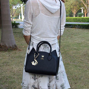 A model carries the black cat-ear handbag on its long shoulder strap outdoors on a path, with the gold-tone crescent and hanging charm visible.
