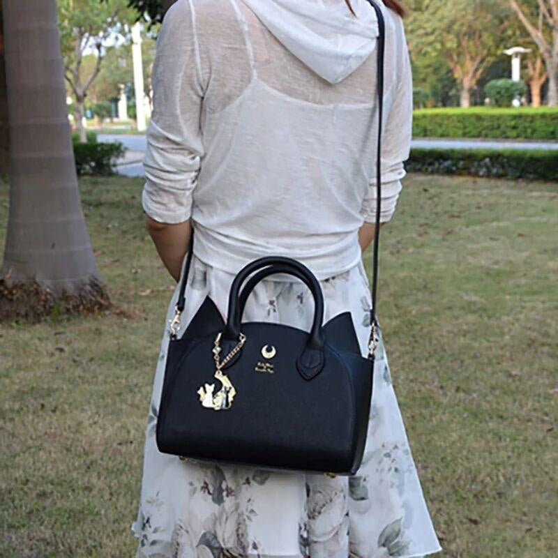 A model carries the black cat-ear handbag on its long shoulder strap outdoors on a path, with the gold-tone crescent and hanging charm visible.