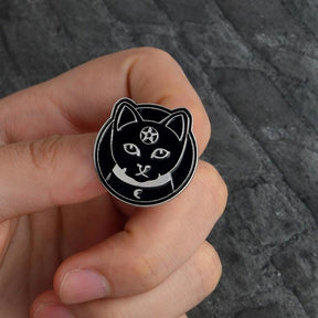 Black cat-face pin held between fingers, showing the pentacle forehead, pointed ears and crescent neck detail.