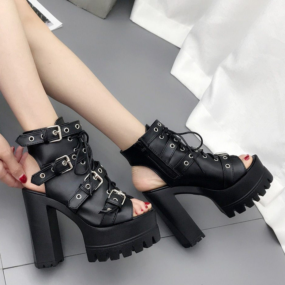 Black buckle booties worn with a white shirt, with one foot lifted to show the open heel area, thick platform and tall block heel.