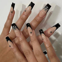 Beige press-on nails with long square black French tips, small silver-tone studs and raised flower-like ornaments, shown across two hands.