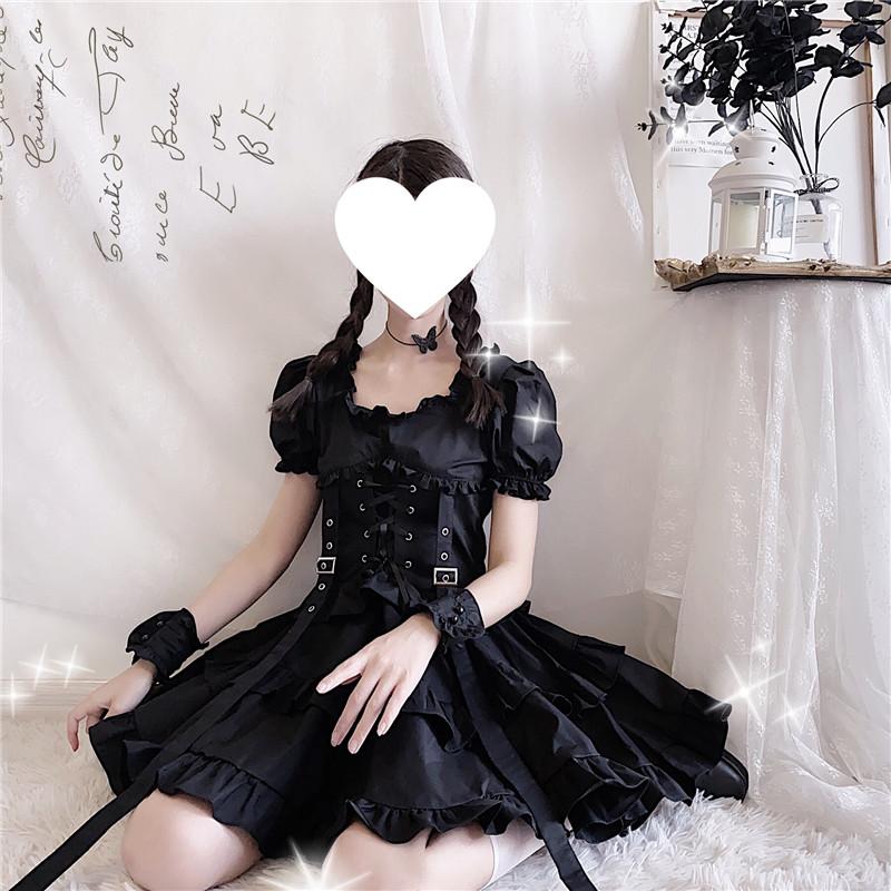 Black gothic ruffle dress worn seated against a pale wall, with the skirt spread wide and the corset harness centred at the waist.