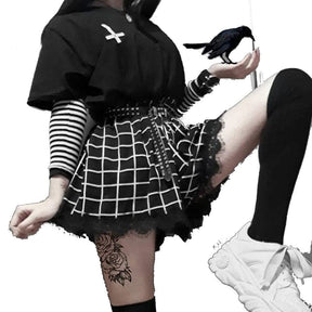The black grid-print skort shown in a raised-leg pose against white, with a striped sleeve, studded belt and thigh-high sock framing its flared lace-trimmed shape.