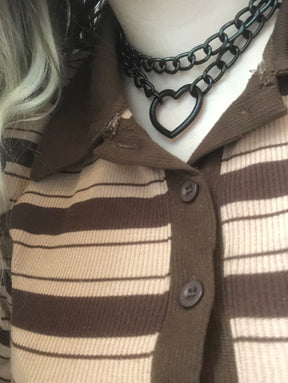 Black heart chain choker worn above a brown striped collared top, showing the two rows of links and hanging open heart against the neckline.