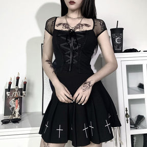 Fuller outfit view of the black lace-up crop top and cross-print skirt, with the model’s hands clasped in front and white cabinets behind.